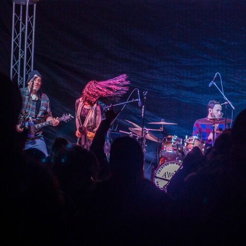 Photo of rock band with lead singer swinging long hair while guitar player and drummer perform.