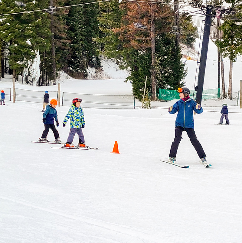 Ski instructor leading two young skiers down the slope around an orange cone