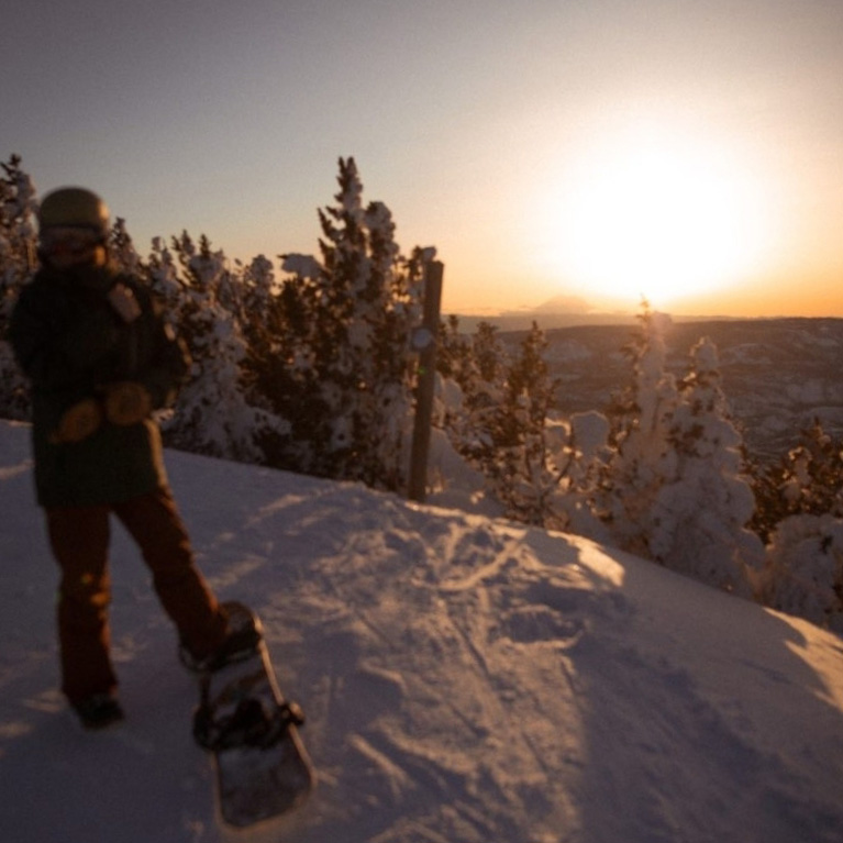 Snowboarder with one foot unstrapped watching sunset behind Mt. Rainier.