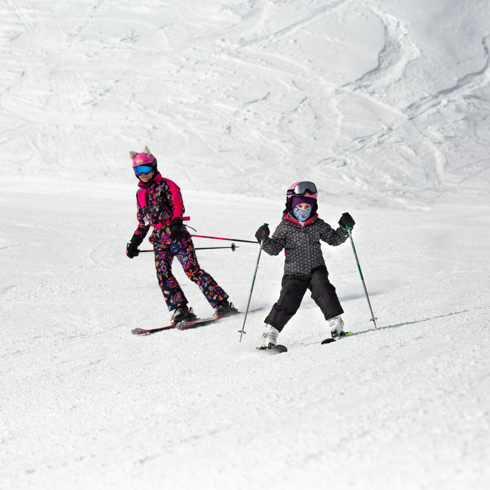 Mother and daughter ski down Lower Bomber Bowl in the sun. Mom is wearing pink and decorative one-piece suite and cat ears on her helmet.