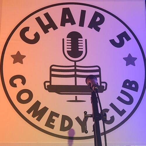 Chair 5 Comedy Club sign with microphone in front casting shadow on sign.