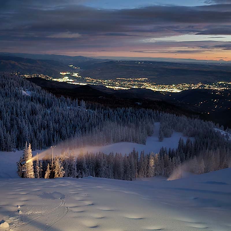 Tower snowgun making snow at night with city lights in the valley below