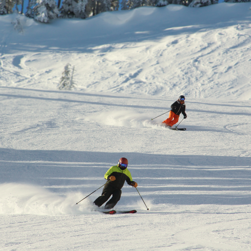 Two skiers carve on a sunny day.