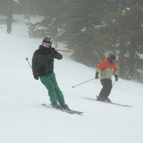 two skiers descend a foggy slope. one greets the camera.