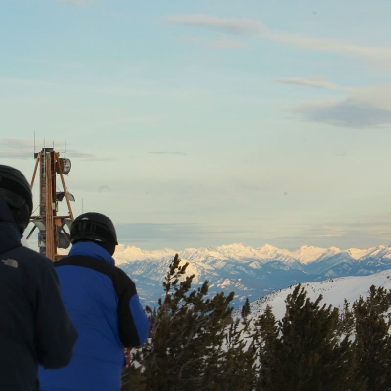 Two men stand in front of the camera. A tower and mountain range are in focus in the distance.