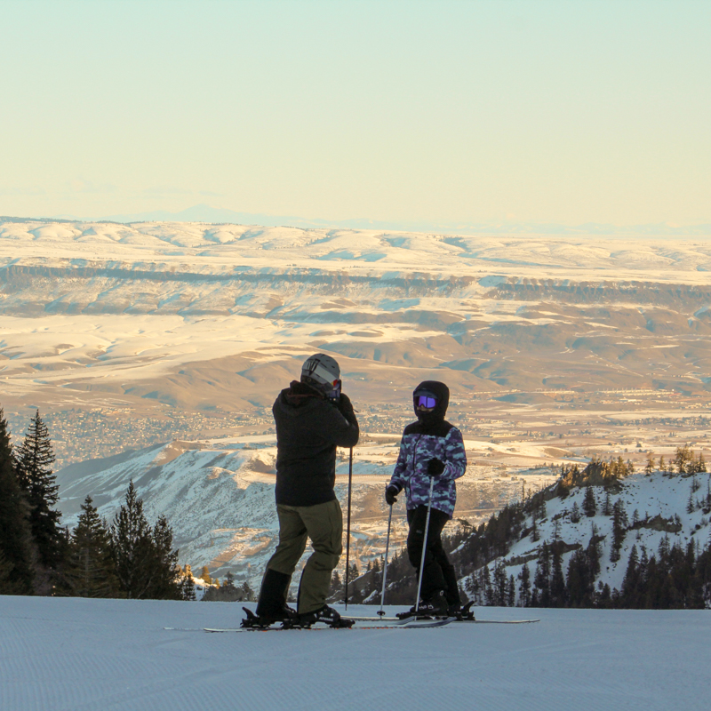 A father and child stop for a photo on a ski slope in front of a sunny valley