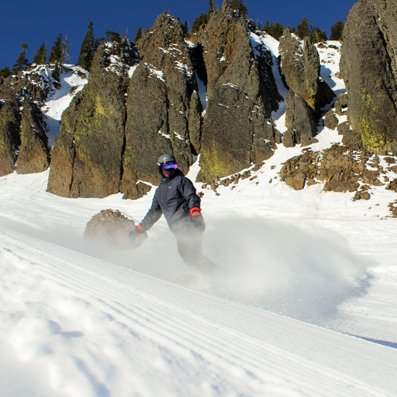 a snowboarder sends up a powder spray in front of basalt cliffs on a sunny day