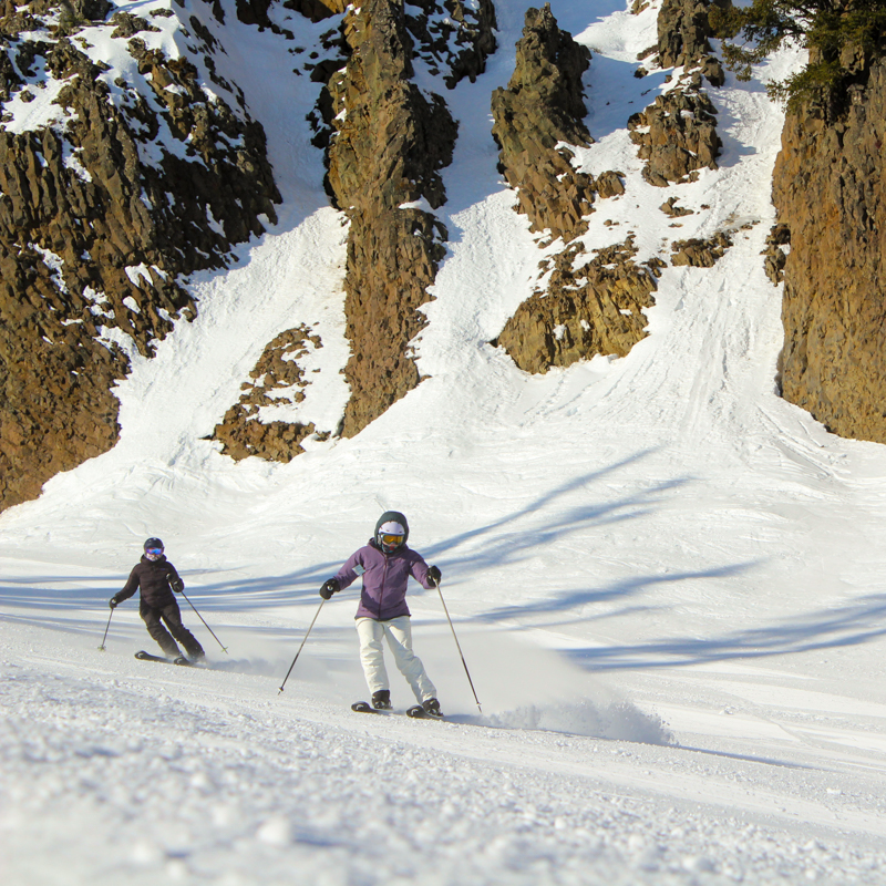 Two woman ski past sunny basalt cliffs