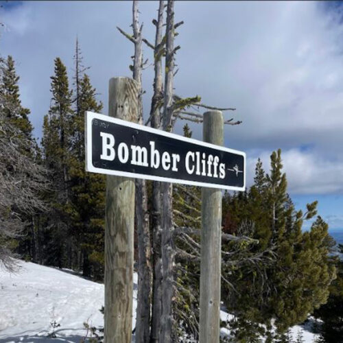 black and white sign reading "Bomber Cliffs" mounted on two wooden posts in a snowy, mountainous landscape. The sign has a small airplane icon on the right side. Behind it, there are evergreen trees, a partly cloudy sky, and snow-covered ground