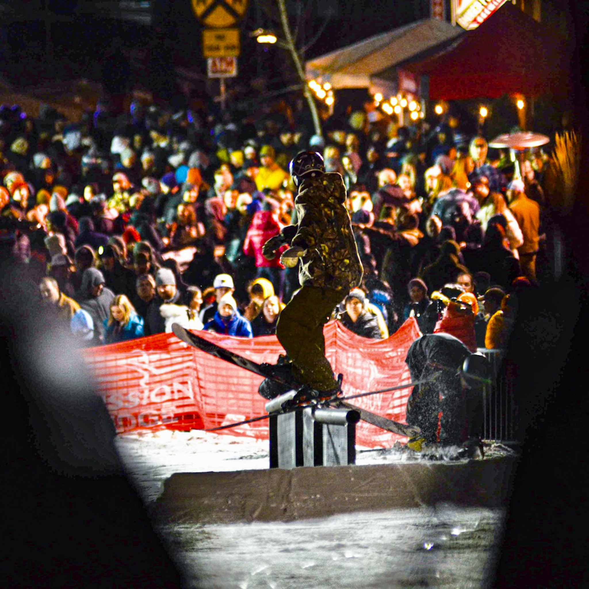 A vibrant nighttime scene capturing a skier performing a rail slide during a lively outdoor event. The rider, dressed in a camo outfit, is illuminated by bright event lighting, with a large crowd of spectators in the background creating an energetic and festive atmosphere. Red safety fencing and event branding add to the organized feel of the competition, while snow and action details highlight the dynamic moment.