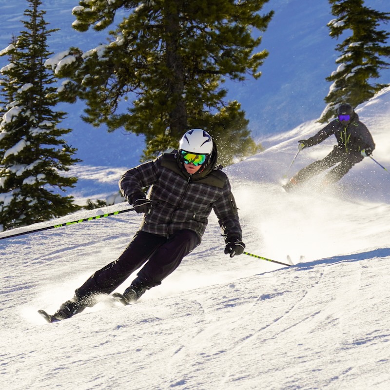 A skier carves a turn on the Bomber Bowl trail at Mission Ridge on a sunny day. Another skier is in the background also making a turn.