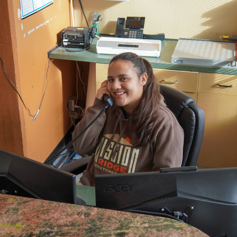 A friendly scene featuring a smiling staff member seated at a desk, speaking on the phone. She is wearing a Mission Ridge hoodie, creating a welcoming and professional impression. The workspace includes dual monitors, office equipment, and a warm-toned background, emphasizing a productive and approachable environment.