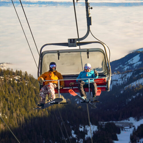 A snowboarder and a skier ride Chair 2 at Mission Ridge on a sunny morning. The skier is smiling and doing a thumbs up.