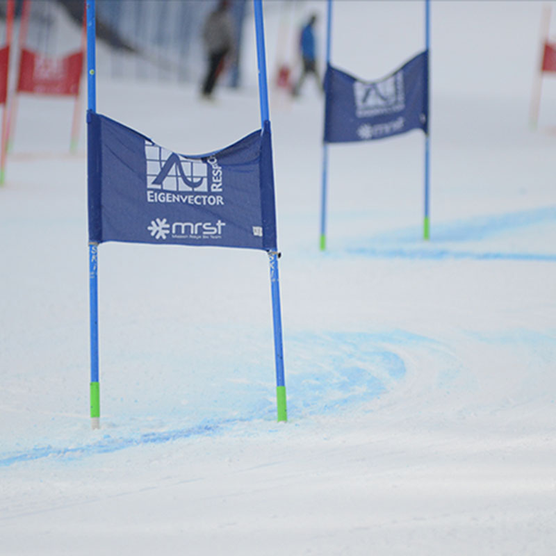 A close-up shot of a ski racing course gate featuring a blue banner with the "Eigenvector Research" logo and the "MRST" (Mission Ridge Ski Team) logo. The gate poles are bright blue with green tips, set against a groomed snow surface with faint blue dye marking the course. The background shows additional gates and blurred figures, emphasizing the action and preparation of a ski racing event.