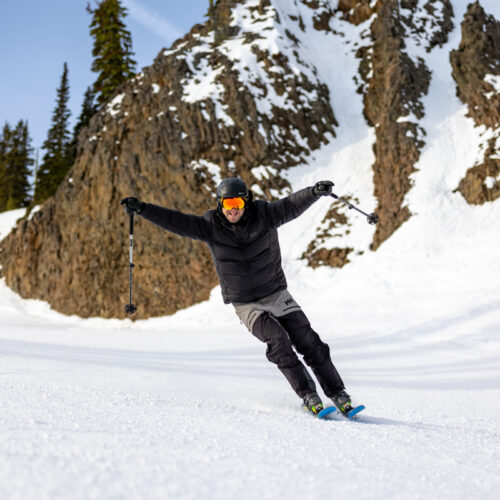 A male skier raises his arms and caws like a bird in front of snowy basalt.