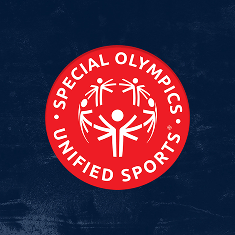 The logo of Special Olympics Unified Sports features a vibrant red circular design with white text reading "Special Olympics Unified Sports" around the border. At the center, stylized figures join hands in a unified, celebratory pose, symbolizing inclusion and teamwork. The navy blue textured background provides a striking contrast, emphasizing the logo's message of unity and sportsmanship.