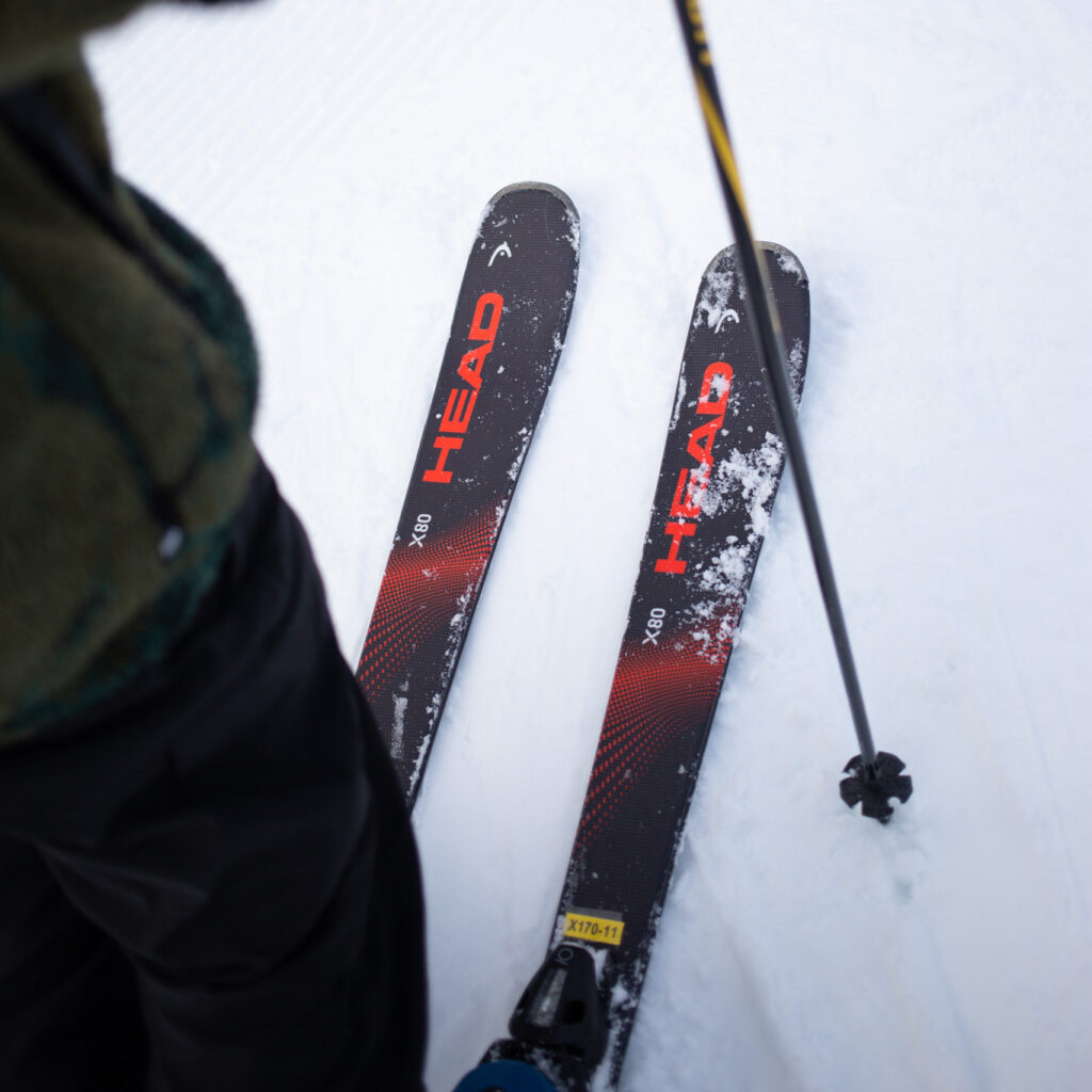 A dynamic close-up of a skier's HEAD X80 skis resting on freshly groomed snow. The vibrant red branding contrasts against the black skis, with patches of snow adding a sense of action and use. A ski pole and part of the skier's outfit are visible, emphasizing the moment's active and authentic nature.