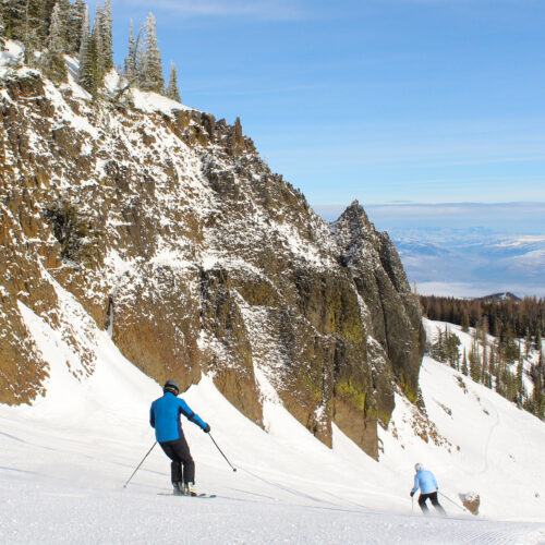 a pair of skiers descend past basalt cliffs on a sunny day