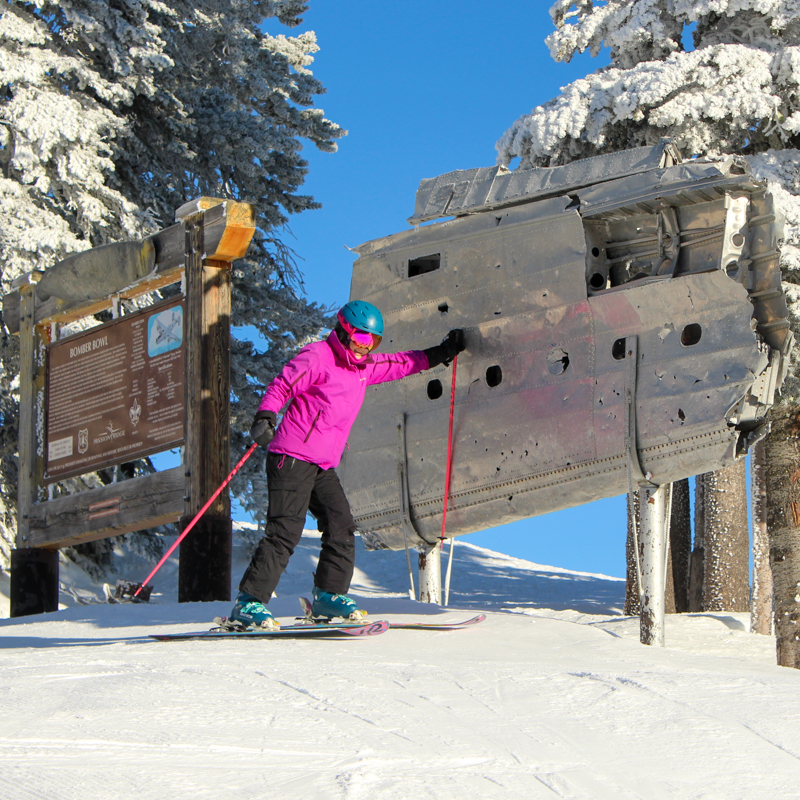 A skier in pink touches a bomber wing monument in front of rimey trees and a clear blue sky.