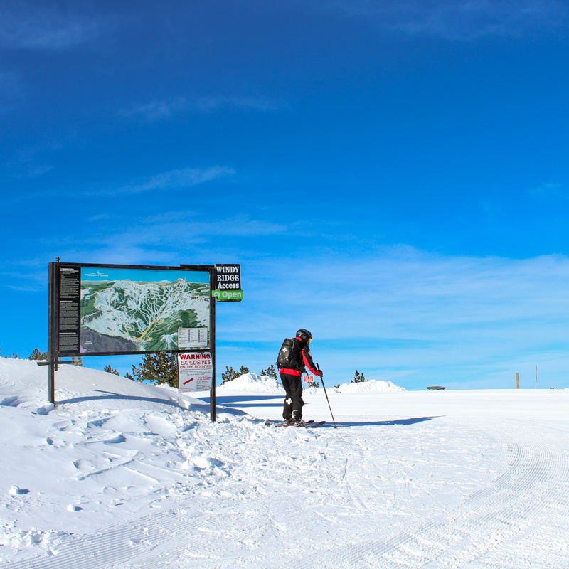 a patroller adjusts his skis in front of a large map on a sunny day