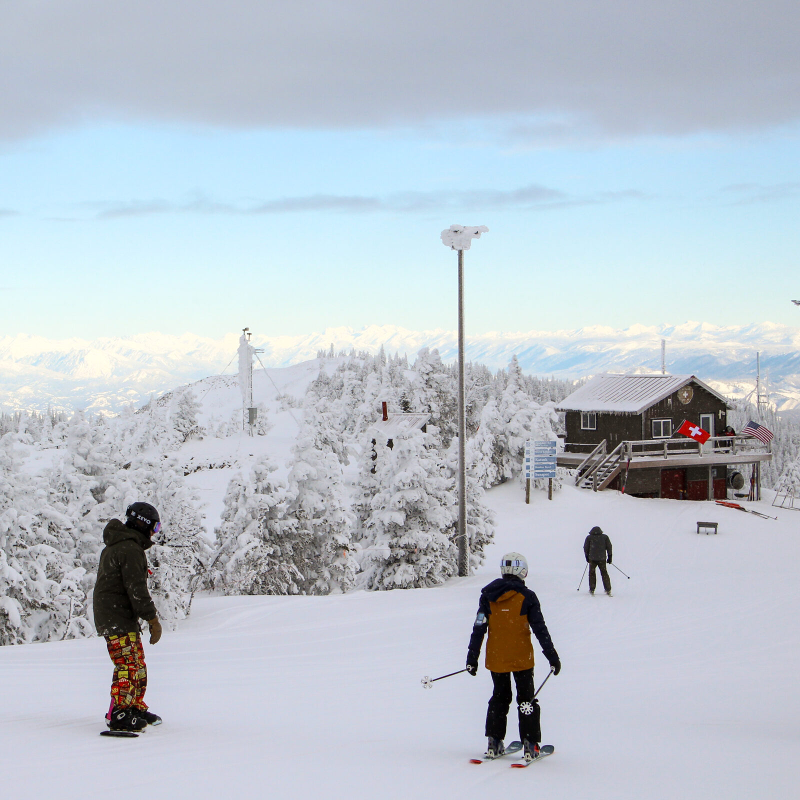 A family of three prepares to ski and board down a fresh snowy mountain on a partly cloudy day. A patrol shack and light posts sit between rimey conifers in the background,