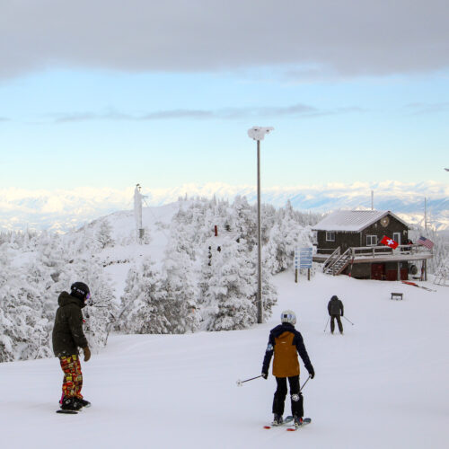 A family of three prepares to ski and board down a fresh snowy mountain on a partly cloudy day. A patrol shack and light posts sit between rimey conifers in the background,