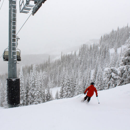 a young skier in red drops into a run along a chair lift tower, with snowy trees and foggy cliffs in the background.