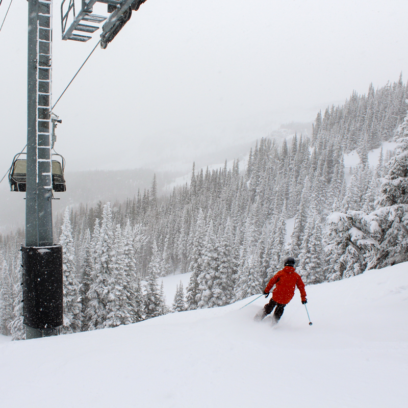 a young skier in red drops into a run along a chair lift tower, with snowy trees and foggy cliffs in the background.