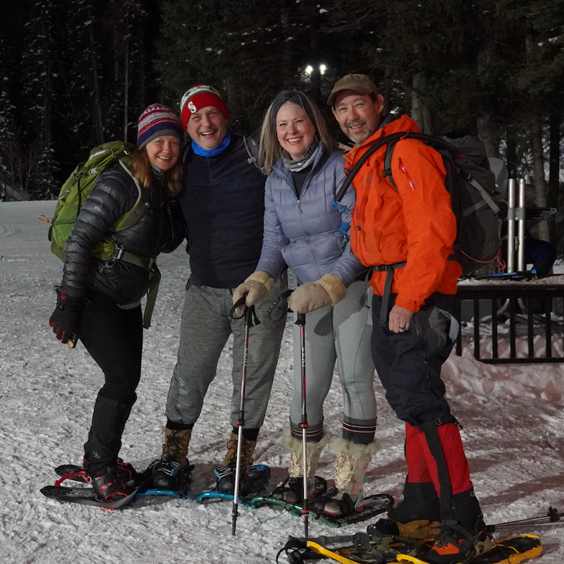 A group of snowshoers smiles for the camera in the night at Mission Ridge, with dark forest and snow behind them.