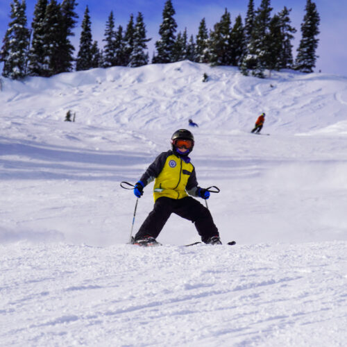 A kid in a yellow jacket descends the Bomber Trail at Mission Ridge on a sunny day.