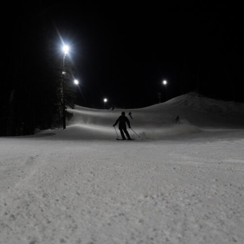 Skiers descend the Tumwater trail at Mission Ridge in the night, with the lights on above.