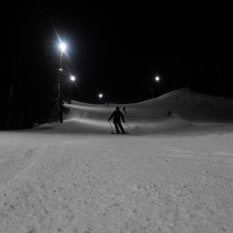 Skiers descend the Tumwater trail at Mission Ridge in the night, with the lights on above.