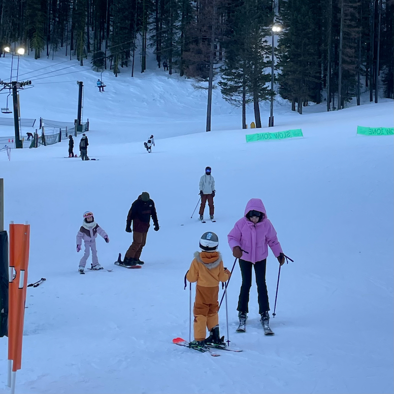 A family chats at the base of the Mimi run at Mission Ridge ski area in the evening with the lights on in the background.