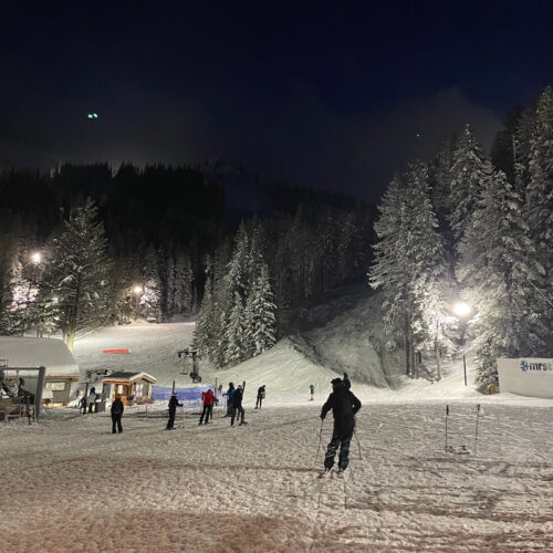 A smattering of skiers and snowboarders stand at the Mission Ridge base area, with some snow covered trees and the night sky in the background.