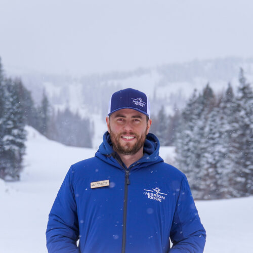 A portrait of new Mission Ridge GM Matt Neubauer standing outdoors in a snowy mountain landscape. He is wearing a blue Mission Ridge jacket and a matching cap, with a name badge visible on his jacket. Snow-covered trees and ski slopes form the background, creating a professional and welcoming winter setting.