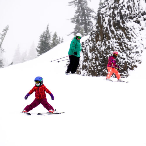 A joyful winter scene featuring three skiers descending a snow-covered slope. In the foreground, a young child in a bright red jacket and purple pants practices skiing in a wedge stance. Behind them, another child in pink and orange ski gear follows with confidence, while an adult in a green jacket observes from above. Snow-covered trees and a rocky outcrop add depth to the scenic alpine setting.