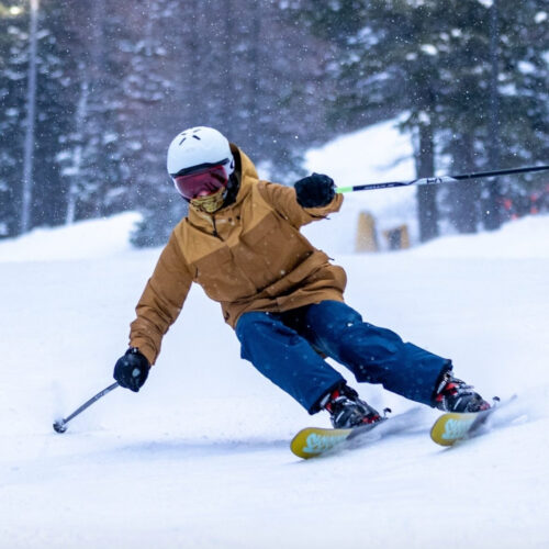 A dynamic action shot of a skier carving aggressively down a groomed slope. Dressed in a tan jacket, blue pants, and a white helmet with red goggles, the skier leans into the turn, kicking up snow with precise edge control. Snowflakes are visible in the air, adding to the wintry atmosphere, while blurred trees in the background provide depth and motion to the scene.