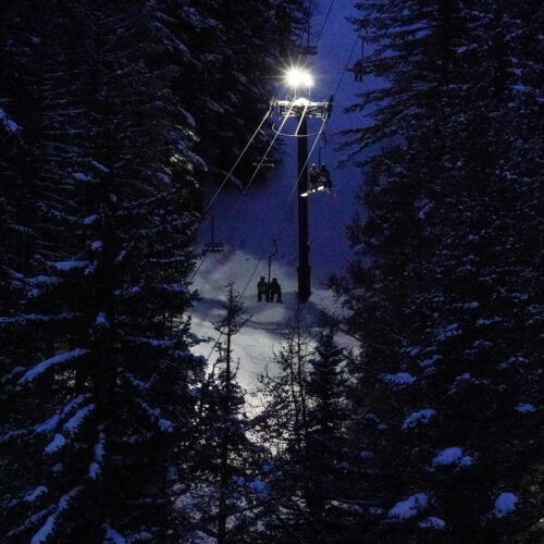 A captivating nighttime scene featuring a ski lift illuminated by bright overhead lights, casting a glow on the snow-covered slope below. The lift ascends through a dense forest of snow-laden trees, creating a peaceful yet adventurous atmosphere. Skiers and riders are seen on the chairs, silhouetted against the light, adding a sense of movement and anticipation to the quiet, wintry night.