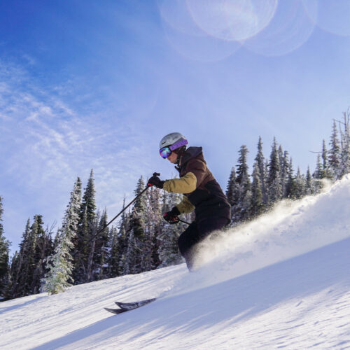 A skier descends a slope at Mission Ridge on a sunny day, with some fresh snow being thrown up behind him.