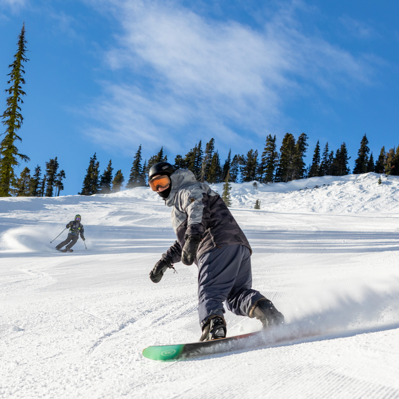 A snowboarder descends the Bomber trail at Mission Ridge on a sunny day, throwing up some snow on a groomed run.