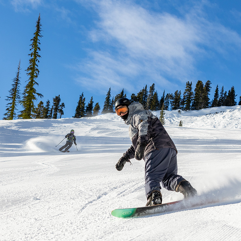 A dynamic action shot of a snowboarder carving through fresh snow, kicking up powder under a bright blue sky. In the background, a skier makes a sharp turn, leaving a trail of snow spray. The sun shines brightly, casting crisp shadows on the well-groomed slope, while tall evergreen trees frame the alpine setting, capturing the excitement of a perfect day on the mountain.