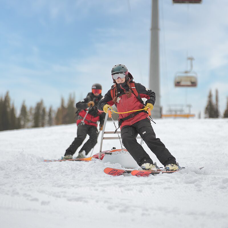 A dynamic action shot of two ski patrollers in red and black uniforms transporting a rescue sled down a snowy slope. The lead patroller skillfully guides the sled with a focused expression, while the second patroller provides support from behind. A ski lift and evergreen trees frame the background, highlighting the alpine setting and the essential work of ski patrol in ensuring mountain safety.